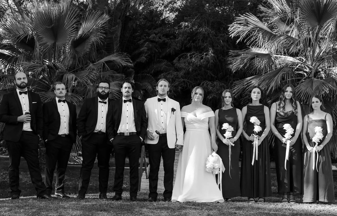 A wedding group photo featuring the bride in a white gown holding a bouquet, the groom in a white tuxedo jacket, and eight bridesmaids and groomsmen in formal attire, standing outdoors in front of large palm trees.