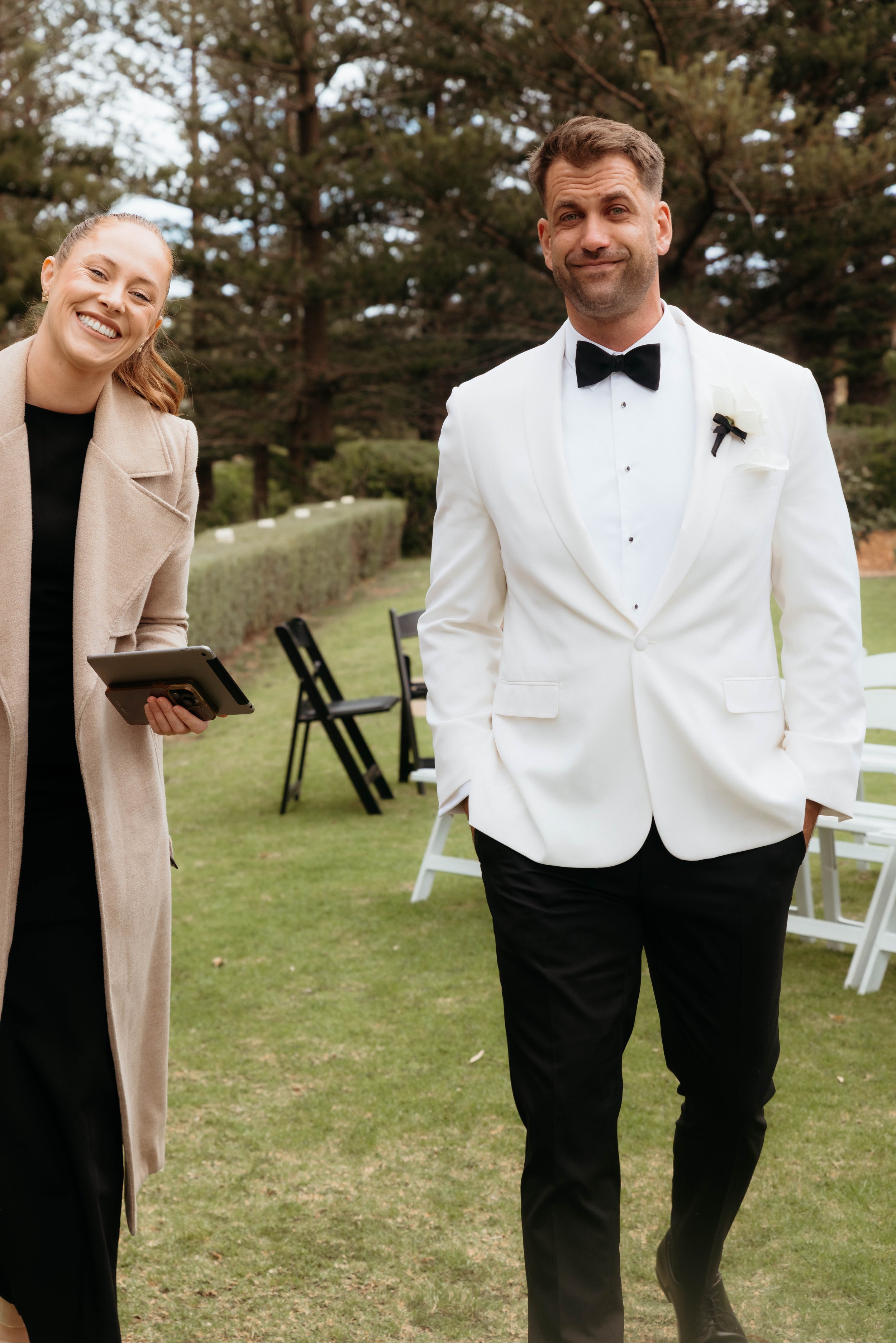 Man in a white tuxedo with a black bow tie and a flower pin, walking outdoors with a woman smiling beside him, outdoors setting with chairs and trees in the background.