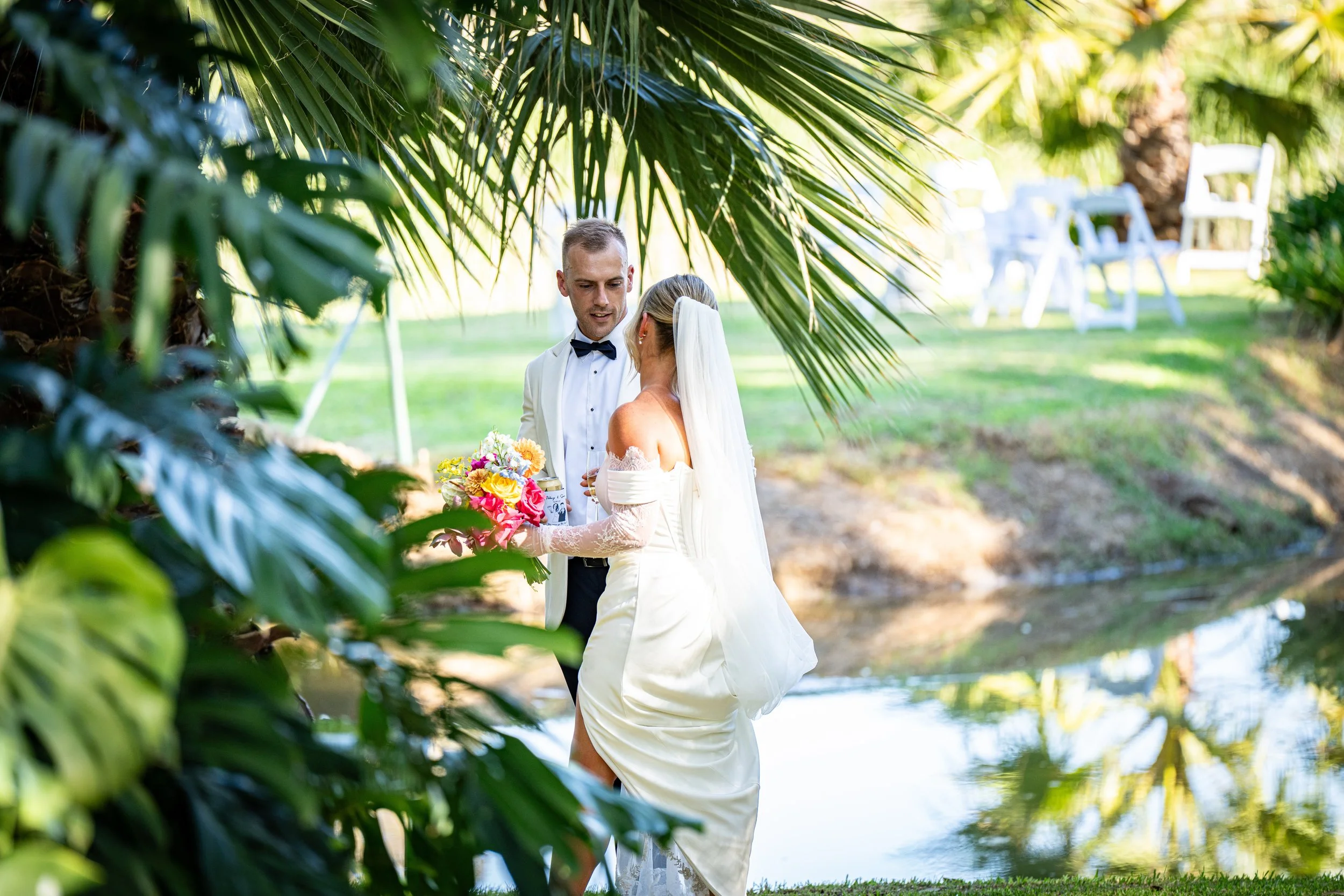 A bride in a white wedding dress and long veil holding a colorful bouquet, standing near a small pond with a groom dressed in a tuxedo with a bow tie, outdoors in a green park setting framed by palm leaves.