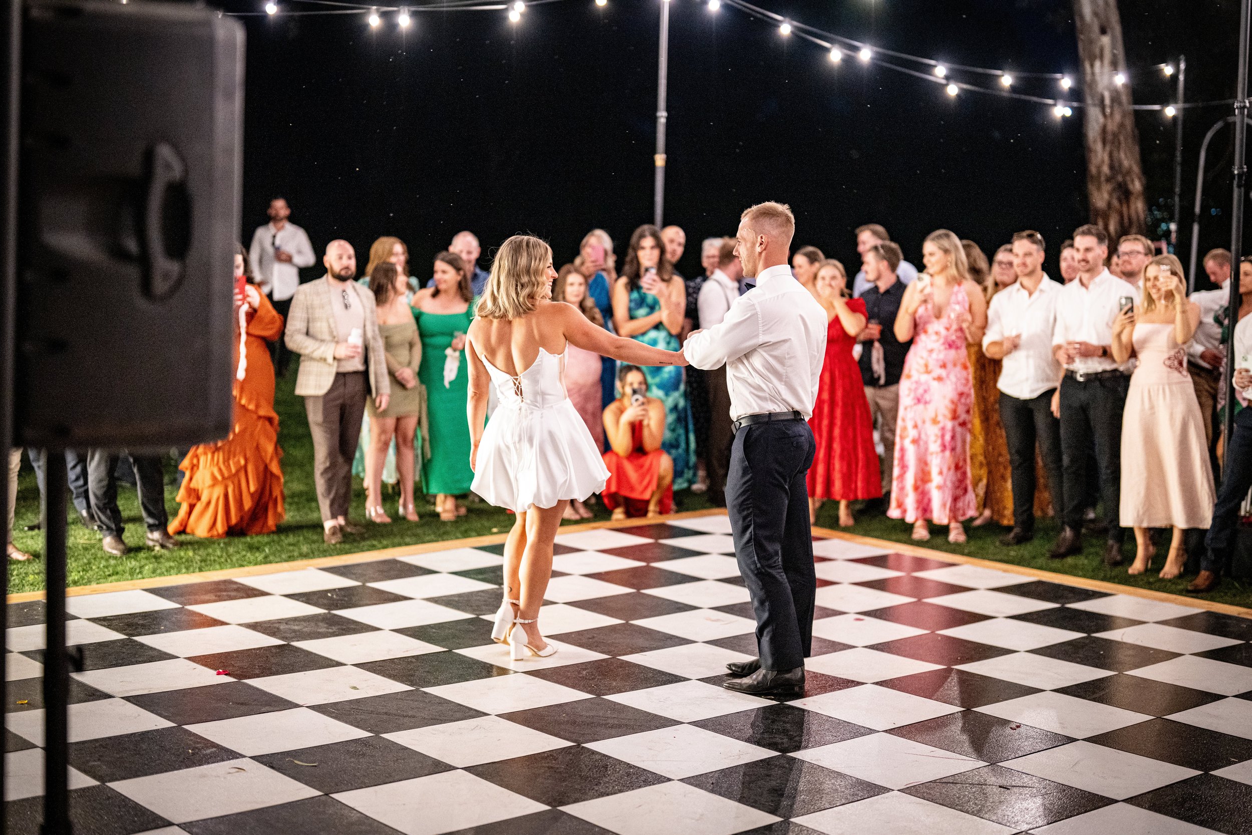 A couple dancing on a checkered dance floor at an outdoor event at night, surrounded by a group of people watching and taking photos.