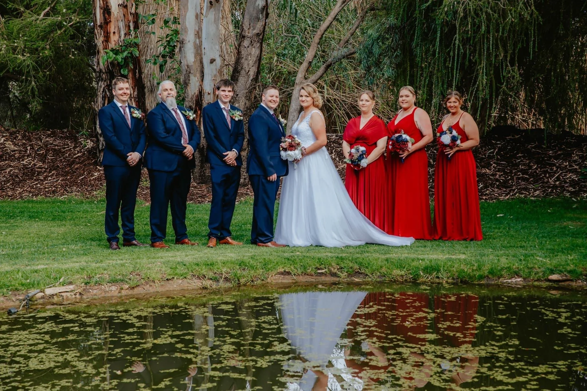 A wedding party standing outdoors near a pond, with a backdrop of trees and greenery. The group includes three men in blue suits, the bride in a white gown, and four women in red dresses, all holding bouquets.