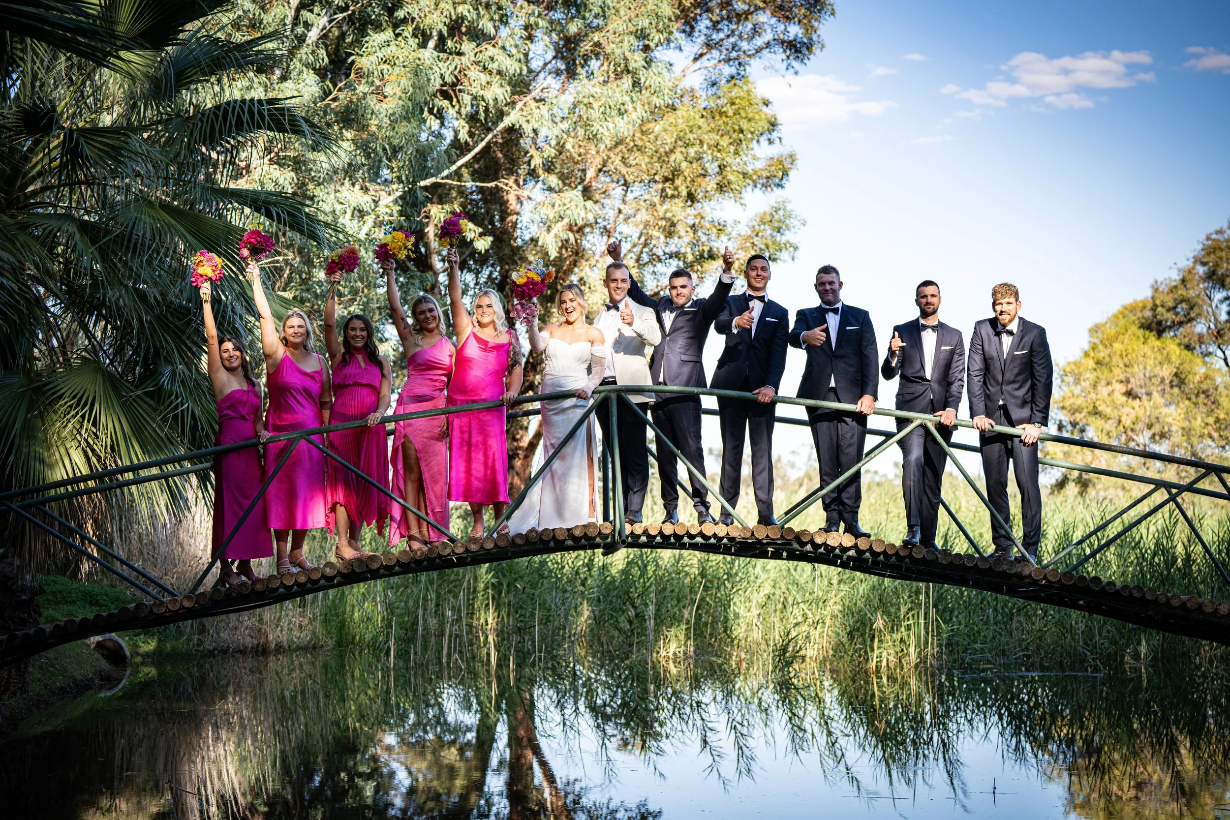A wedding party standing on a small bridge over a pond, with the bride and groom in the center, surrounded by bridesmaids in pink dresses and groomsmen in tuxedos, some holding bouquets and giving thumbs up.