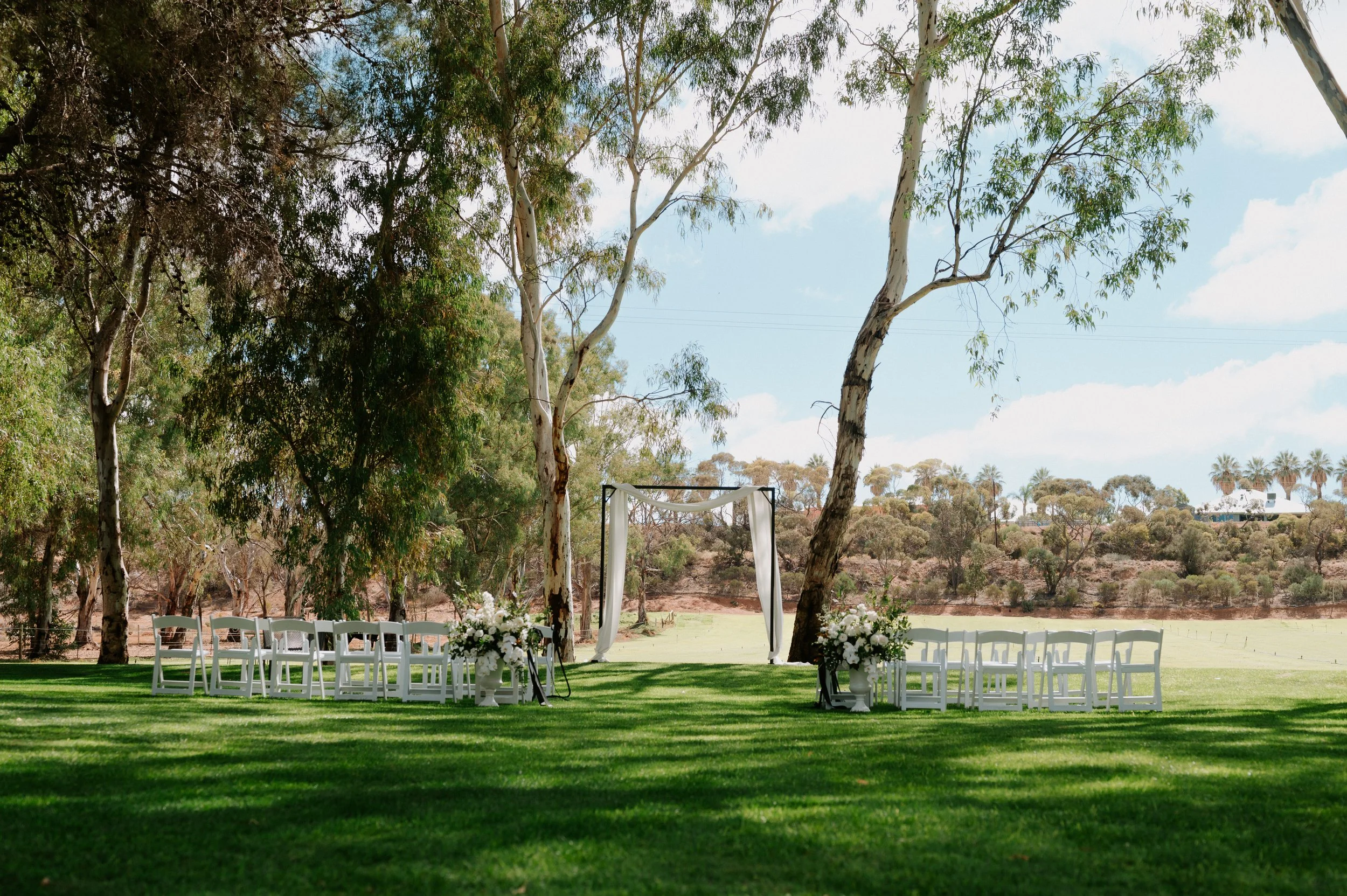 Outdoor wedding setup with white chairs arranged on green grass under tall trees, facing a white fabric-draped archway, with floral arrangements on stands on either side, against a background of a field and scattered trees.