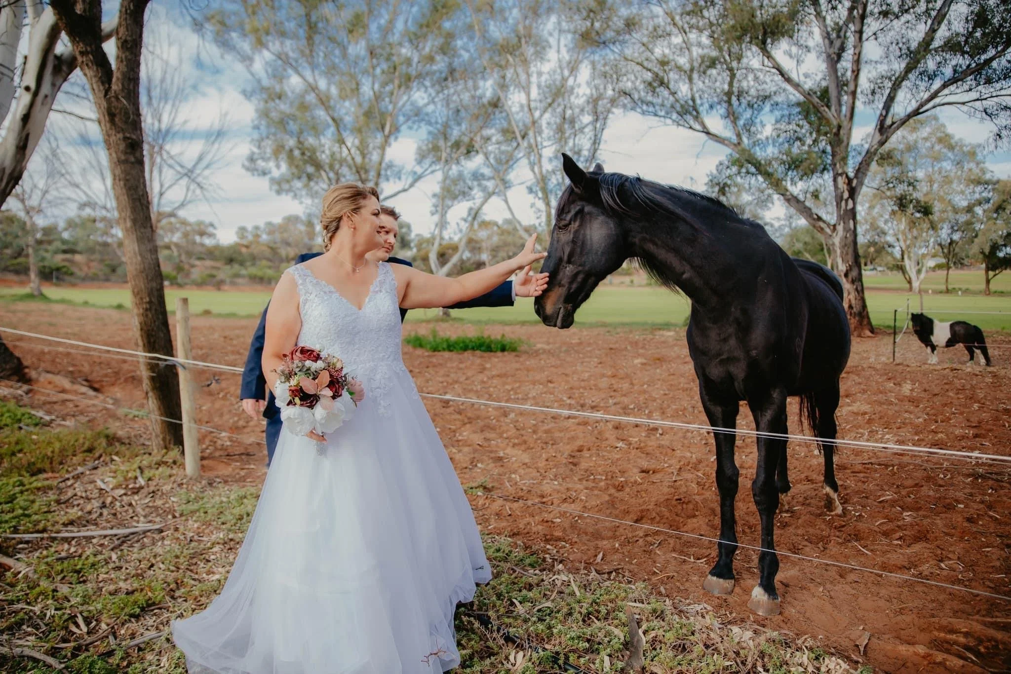 Bride in wedding dress with bouquet petting a black horse in a farm setting with trees and a fence.