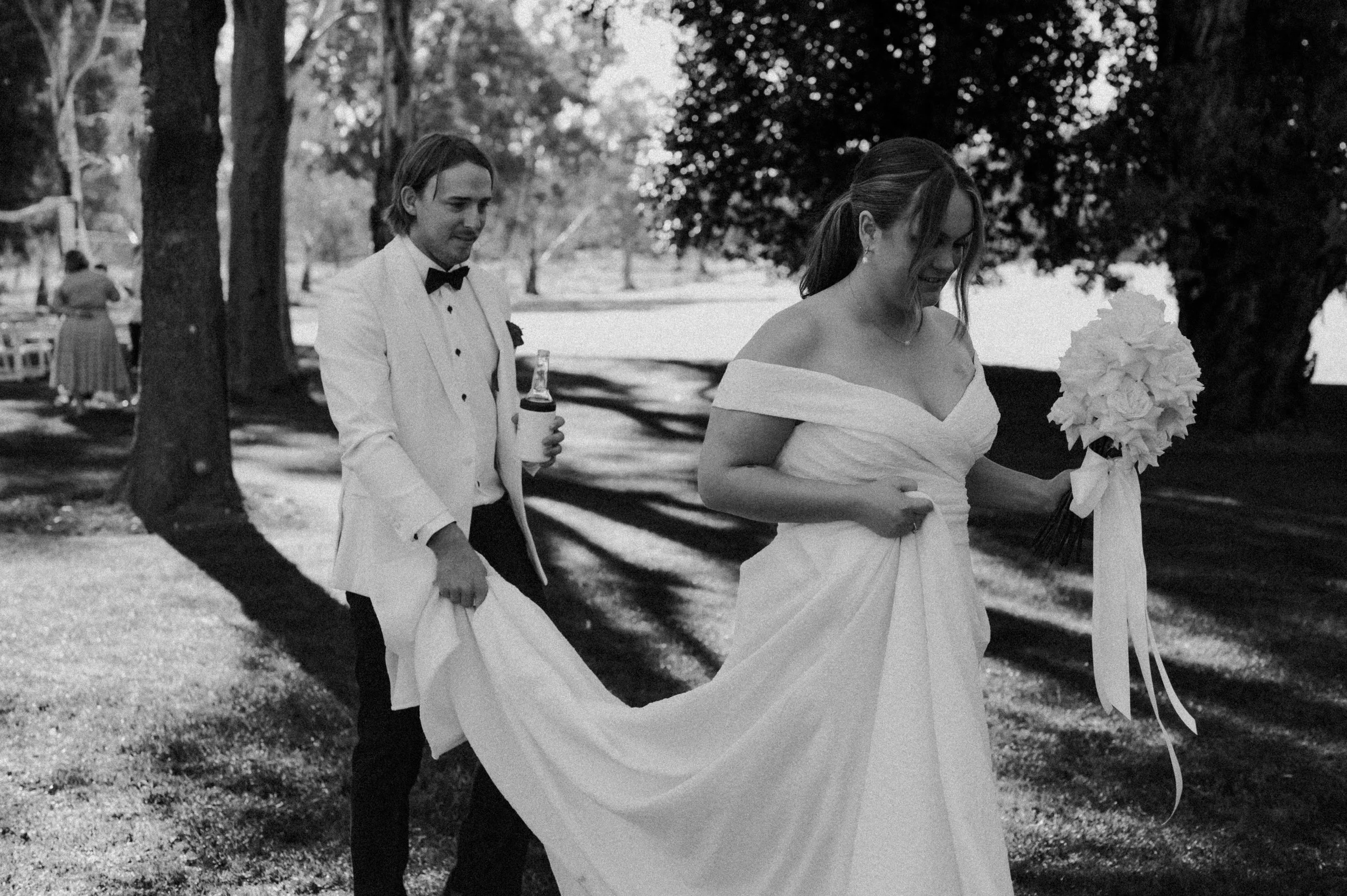A black and white photograph of a bride in an off-the-shoulder wedding gown holding a bouquet, and a groom in a tuxedo with a bow tie, walking outdoors in a park-like setting.