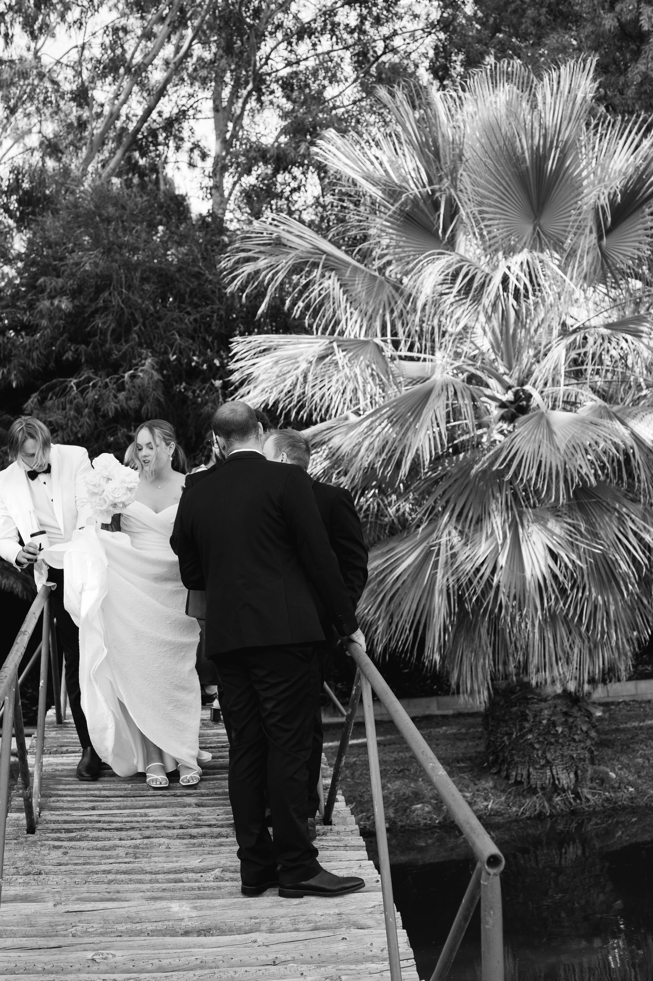 A black-and-white photo of a wedding party on a small wooden bridge outdoors. The bride in a white wedding dress is holding a bouquet, walking with several people, some in formal attire including tuxedos and suits. Large palm tree and lush trees are 