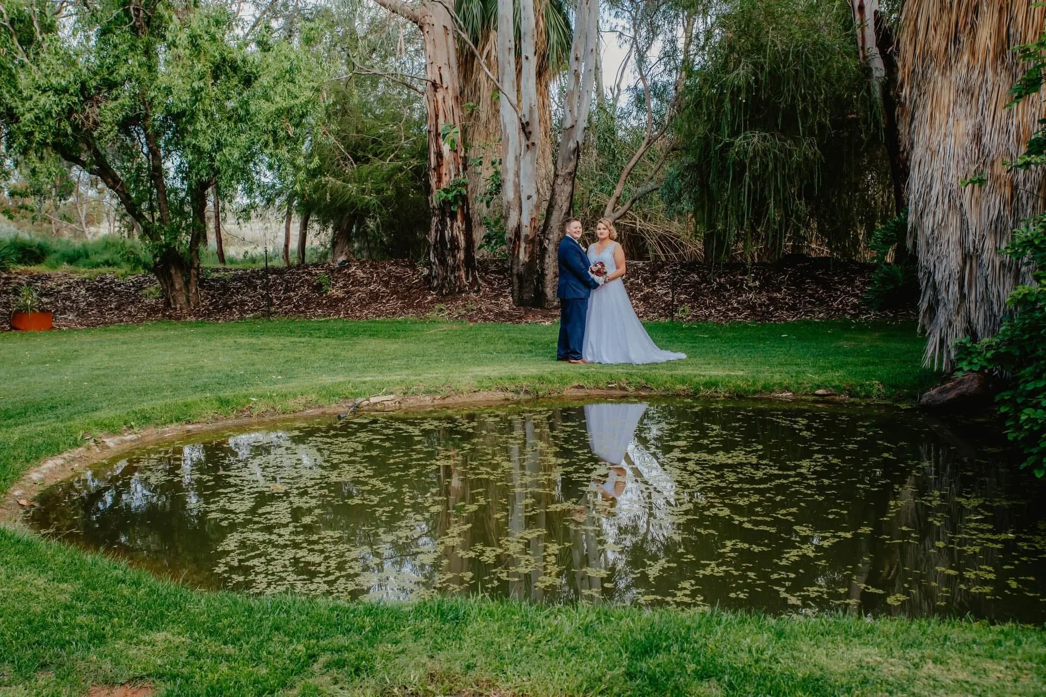 A bride and groom standing near a small pond in a garden, with trees in the background and the pond reflecting their image.
