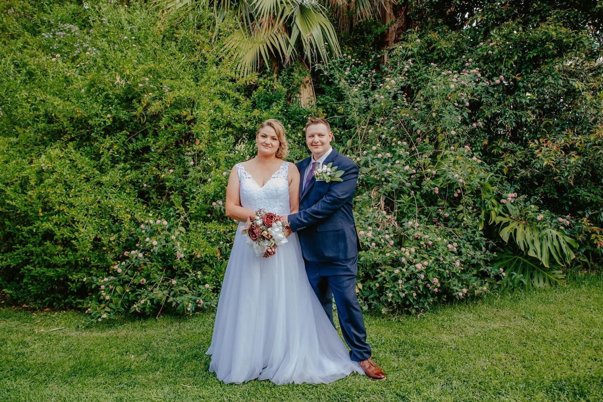A bride and groom stand together outdoors, surrounded by green bushes and plants. The bride wears a white wedding gown and holds a bouquet, while the groom wears a navy suit with a boutonniere.