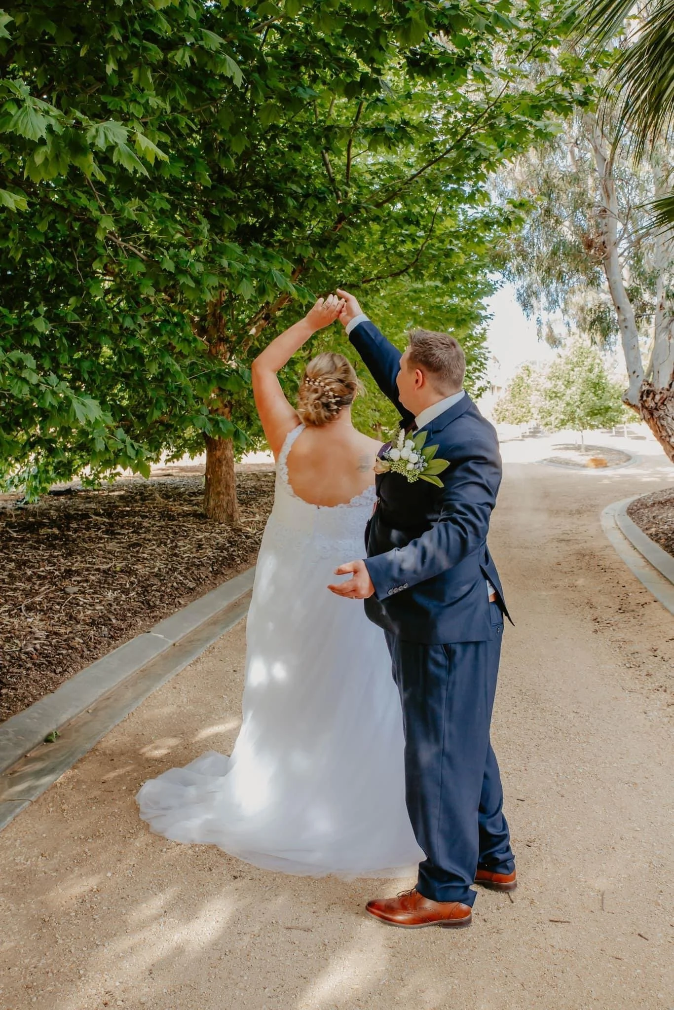 A bride and groom dancing outdoors on a dirt path surrounded by trees with lush green leaves.