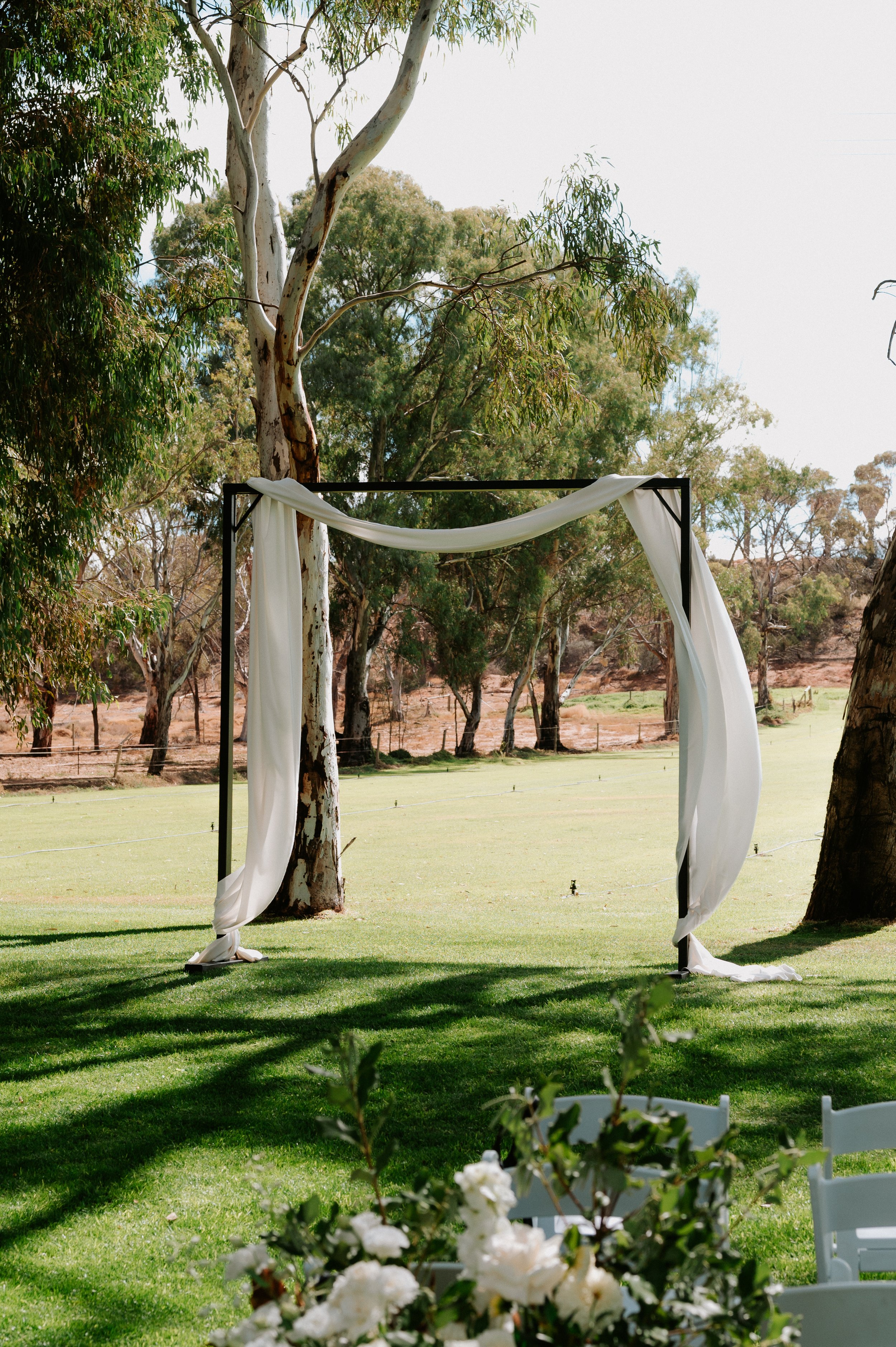 A wedding arch decorated with white fabric on a grassy outdoor area surrounded by trees.
