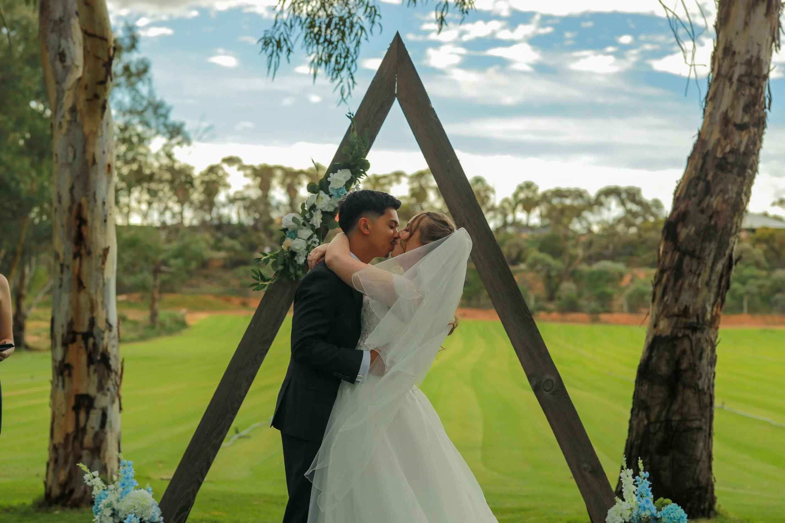 Bride and groom sharing their first kiss beneath a floral wedding arbour during an outdoor ceremony at Greenfield Acres Wedding Venue in the Riverland, South Australia.