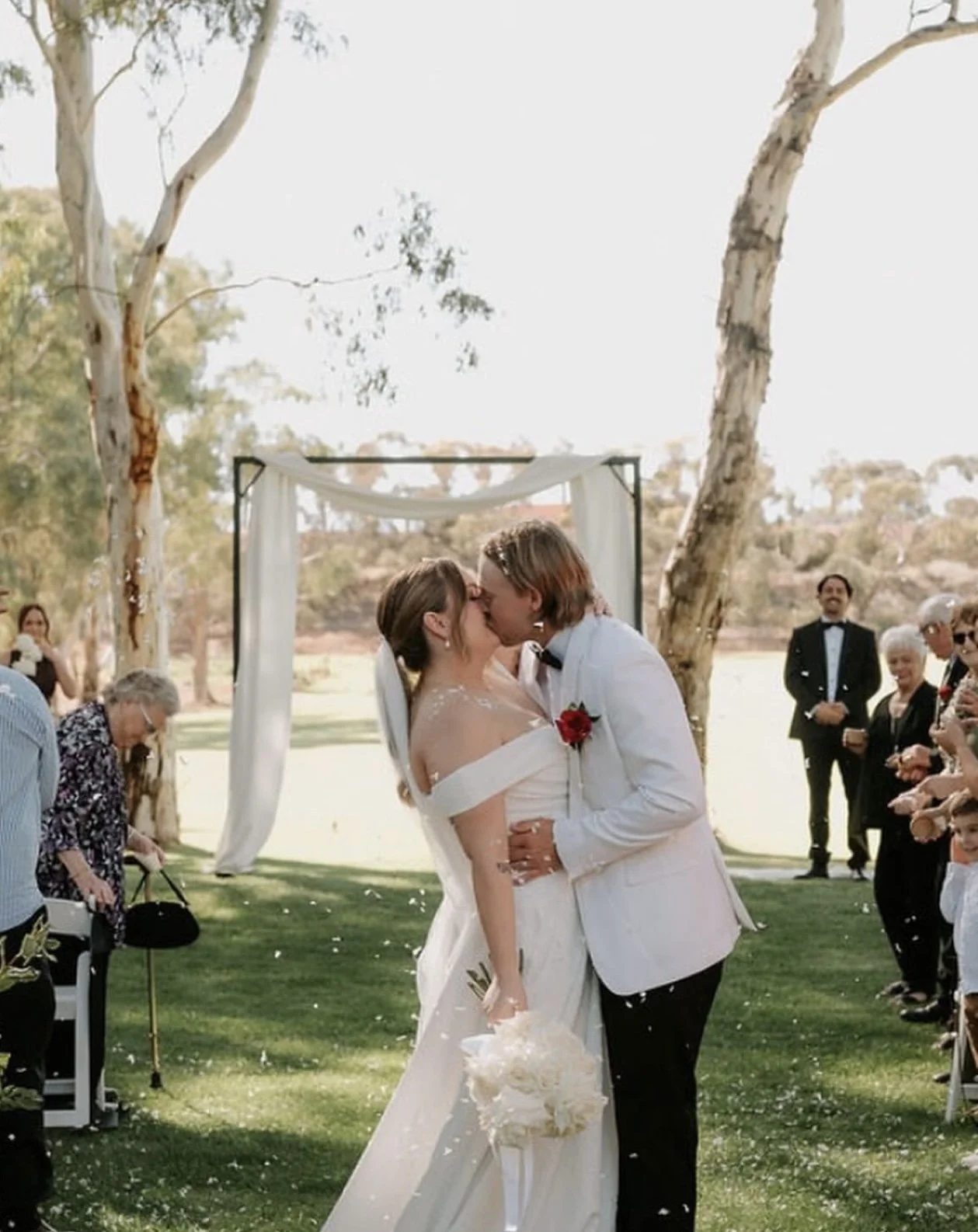 A married couple sharing a kiss at their outdoor wedding ceremony, with guests watching and trees in the background.