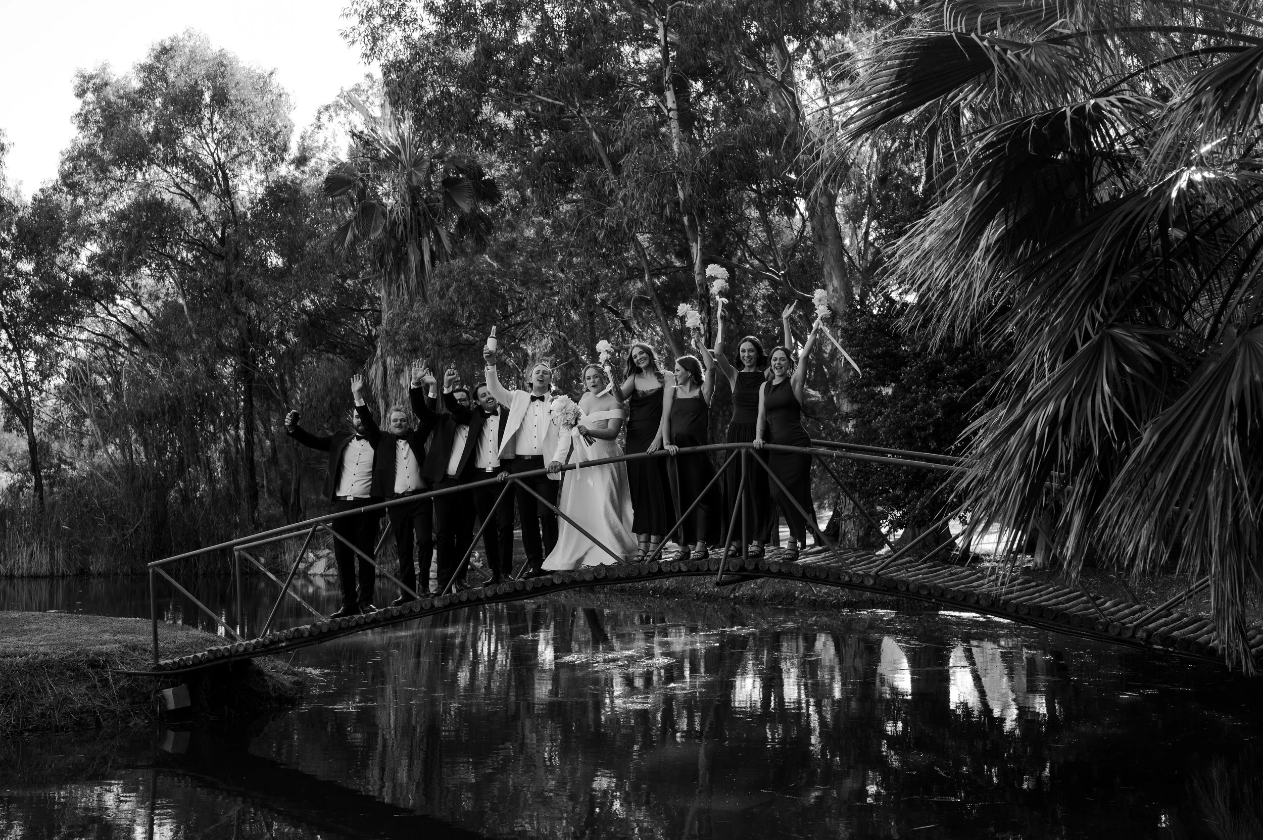 Black and white photo of a wedding party standing on a small bridge over water, with a bride, groom, and bridesmaids holding bouquets and celebrating outdoors surrounded by trees.