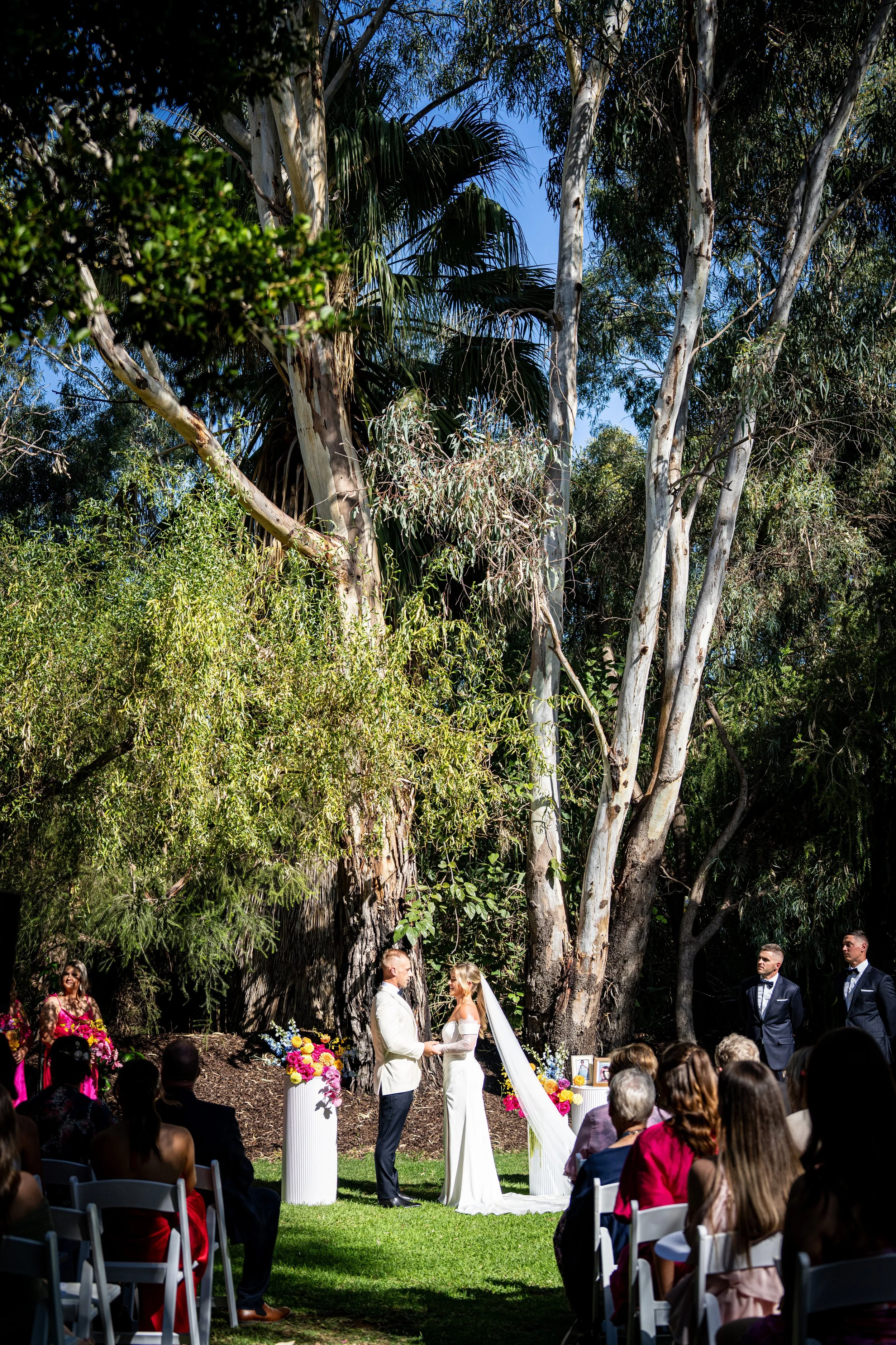 A wedding ceremony outdoors under large trees with a couple holding hands and facing each other, surrounded by bridesmaids and groomsmen, and seated guests watching the event.