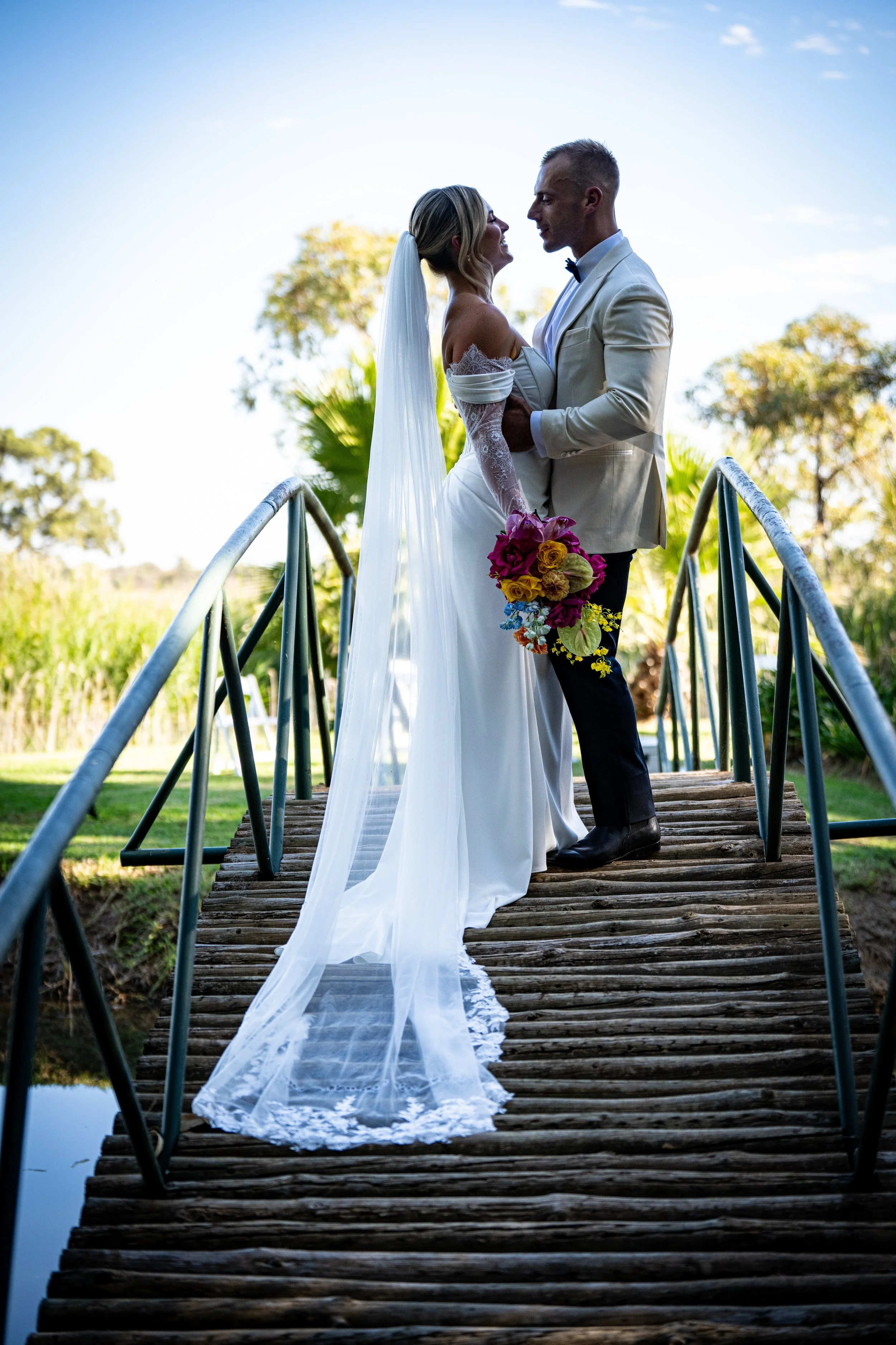 A bride and groom standing on a small wooden bridge outdoors, gazing at each other, with a scenic background of trees and sky.