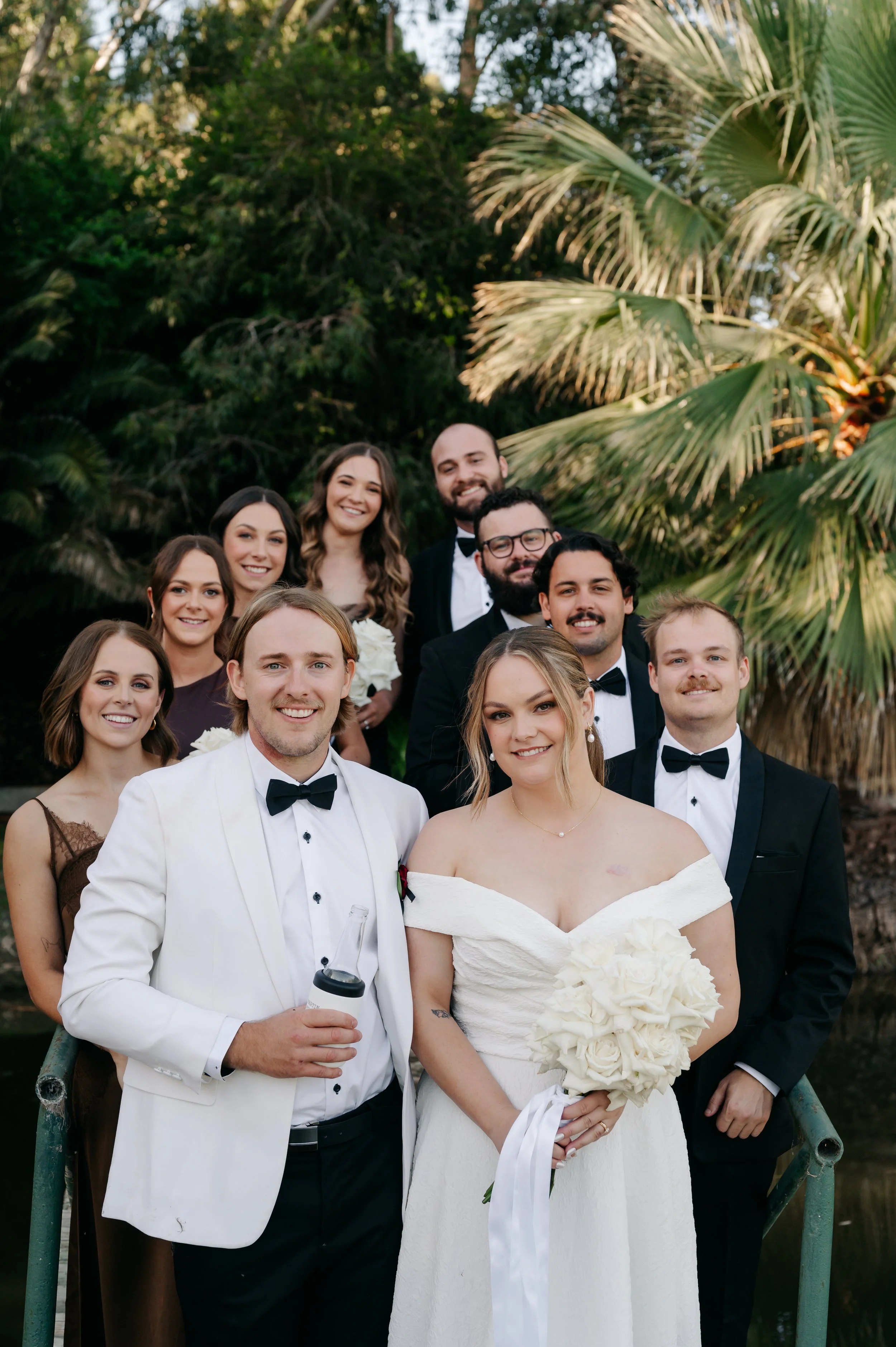 A wedding party of ten people, including the bride in a white off-shoulder gown holding a bouquet of white roses, and the groom in a white tuxedo with a black bow tie, standing outdoors in front of a lush, tropical background.