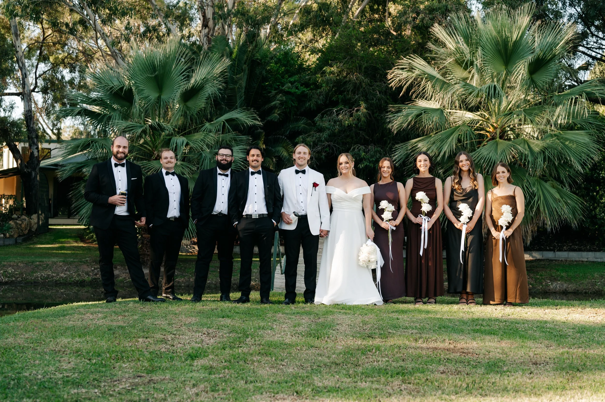 A wedding party of ten people standing outdoors on a grassy lawn, with palm trees and greenery in the background. The group includes groomsmen in black tuxedos, the bride in a white wedding gown and the groom in a white tuxedo jacket, and bridesmaids in dark dresses holding white bouquets.