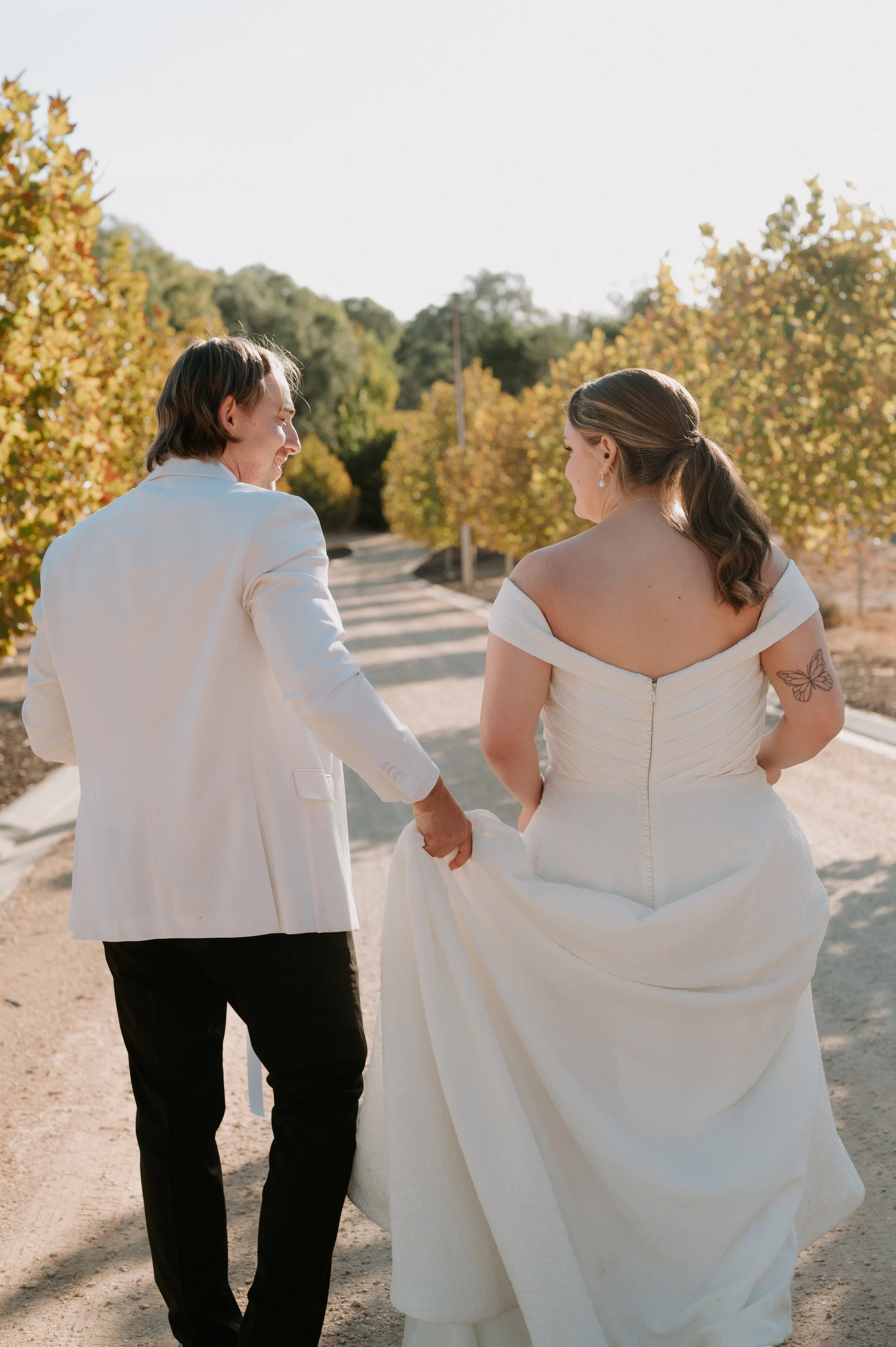 A bride and groom walk hand in hand on a dirt path beside trees with fall foliage, sharing a moment during their wedding.