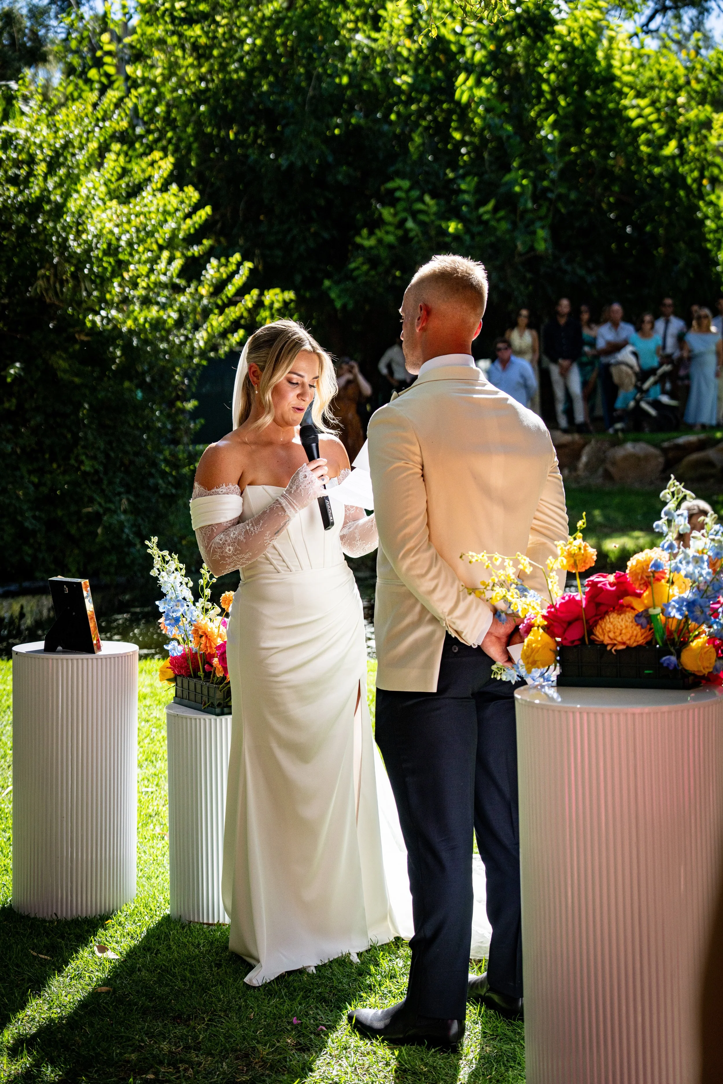 A bride and groom exchange wedding vows outdoors under trees. The bride reads from a paper, holding a microphone, while the groom listens. Guests stand in the background, and colorful flower arrangements decorate the scene.