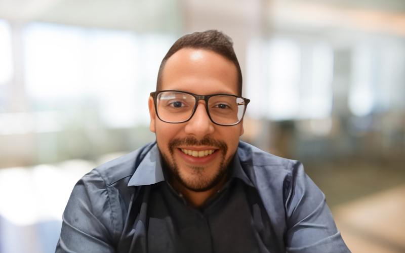 A man with short dark hair, glasses, and a beard smiling at the camera, wearing a dark gray shirt, in an indoor setting with bright natural light.