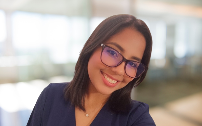 A young woman smiling while taking a selfie in a bright indoor setting, wearing glasses and a navy blue top.