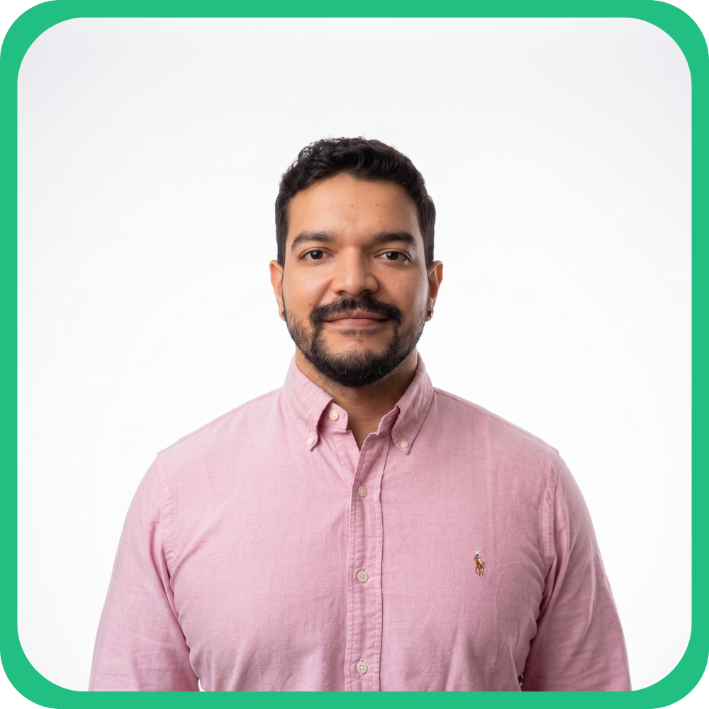A man with dark hair and a beard wearing a pink button-up shirt standing against a white background.