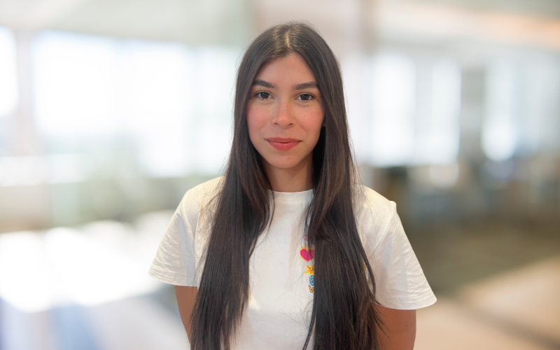 Young woman with long dark hair wearing a white T-shirt standing indoors with a blurred background.