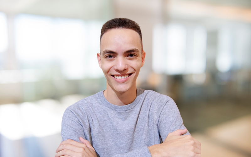 Young man with short hair wearing a gray t-shirt, smiling with arms crossed in a bright indoor setting.