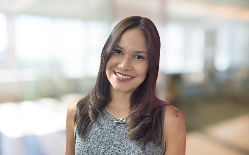 Smiling woman with shoulder-length brown hair, wearing a sleeveless gray top, indoors with blurred windows and light in the background.