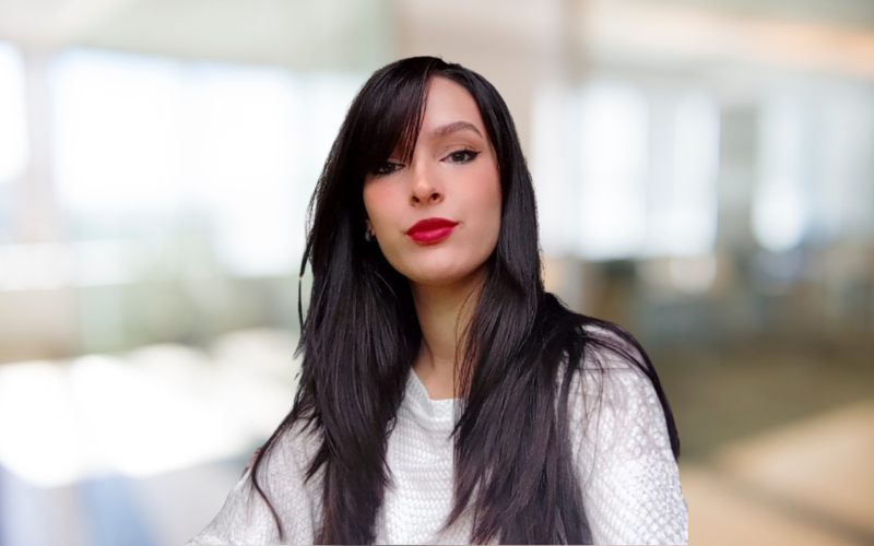 A young woman with long black hair and red lipstick looks at the camera in an indoor setting with blurred windows and light background.