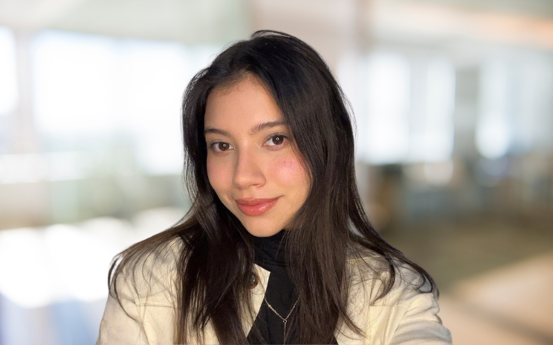 Young woman with long dark hair and light skin, smiling softly, standing indoors with natural light.