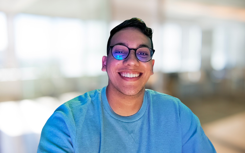 Young man smiling, wearing glasses and a blue shirt, indoors with bright windows in the background.