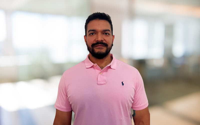 A man with a beard and short dark hair wearing a pink polo shirt standing indoors with a blurred background.