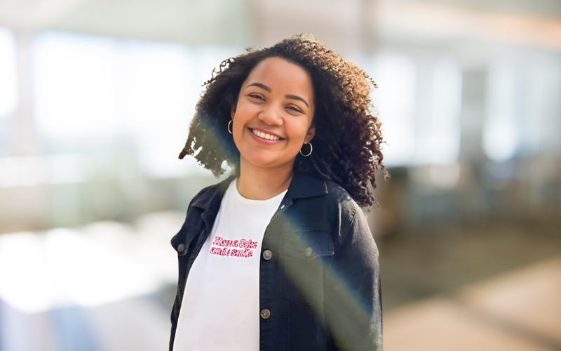 Smiling woman with curly hair wearing a white t-shirt and denim jacket, standing in a bright indoor space.