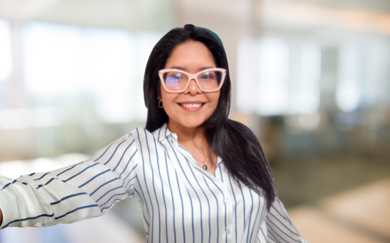 Smiling woman with long dark hair, wearing glasses and a striped shirt, taking a selfie in a bright, modern indoor space.