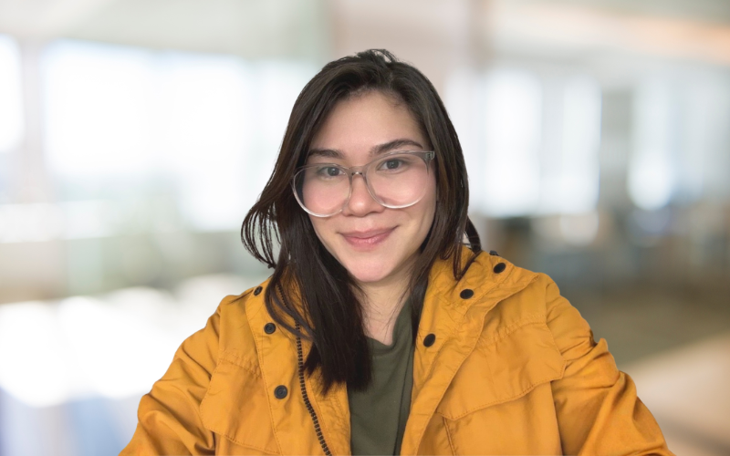 A young woman with glasses and long dark hair, smiling, wearing a yellow jacket indoors.