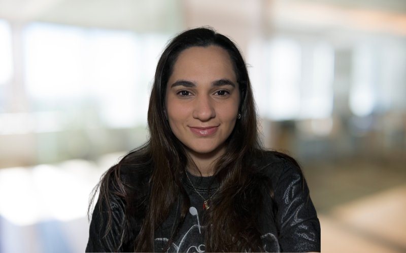 A young woman with long dark hair smiling indoors with a blurred background of windows and furniture.