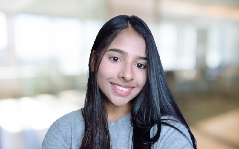 A young woman with long dark hair, smiling and wearing a gray t-shirt in a bright indoor setting.