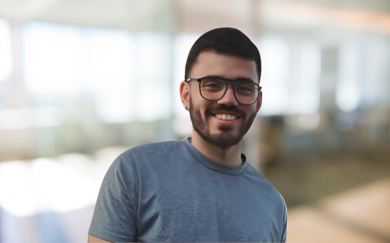 A young man with dark hair, glasses, and a beard smiling in an indoor setting with bright windows in the background.