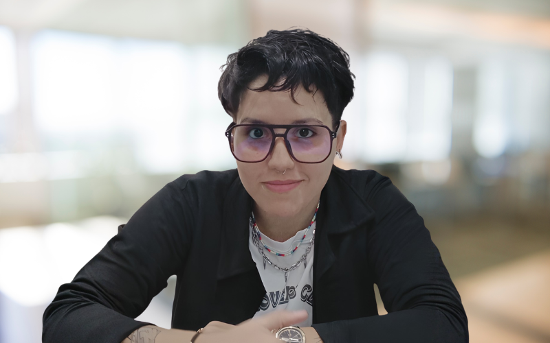A young woman with short dark hair and glasses sitting at a table indoors, smiling at the camera.