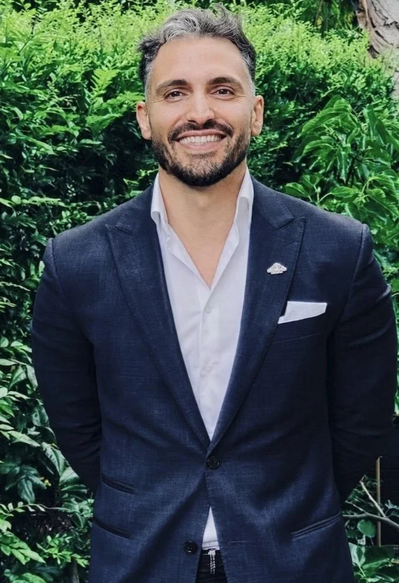 Man in a blue suit standing outdoors with greenery background, smiling.