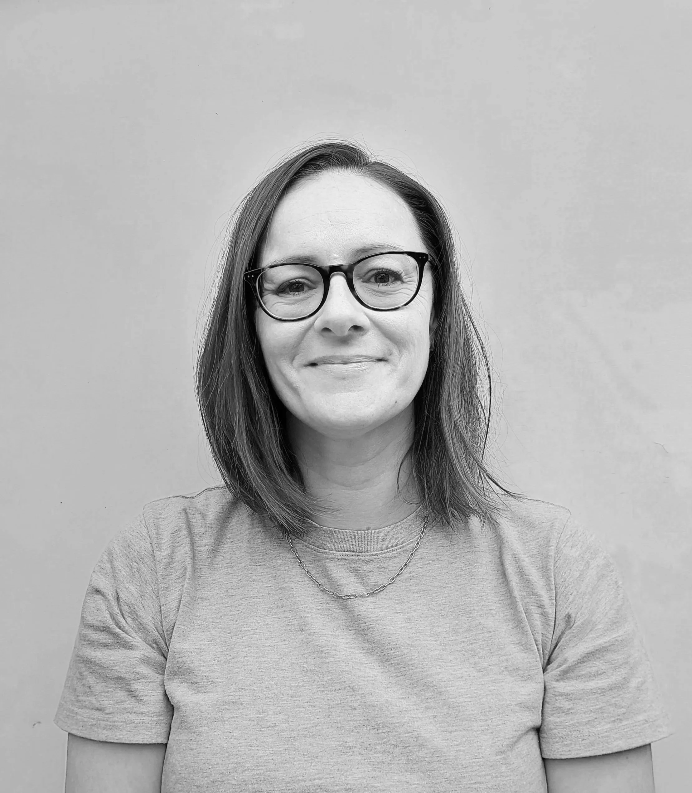 A woman with shoulder-length hair and glasses smiling, wearing a light-colored top and a necklace, standing against a plain wall.
