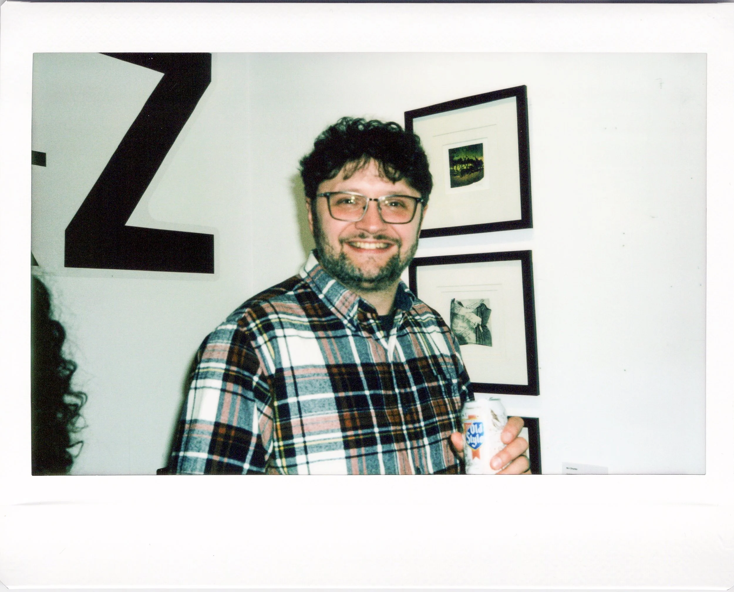 A man with glasses and a beard wearing a plaid shirt, holding a can of Squirt soda, standing in front of a white wall with framed pictures and a large letter Z.