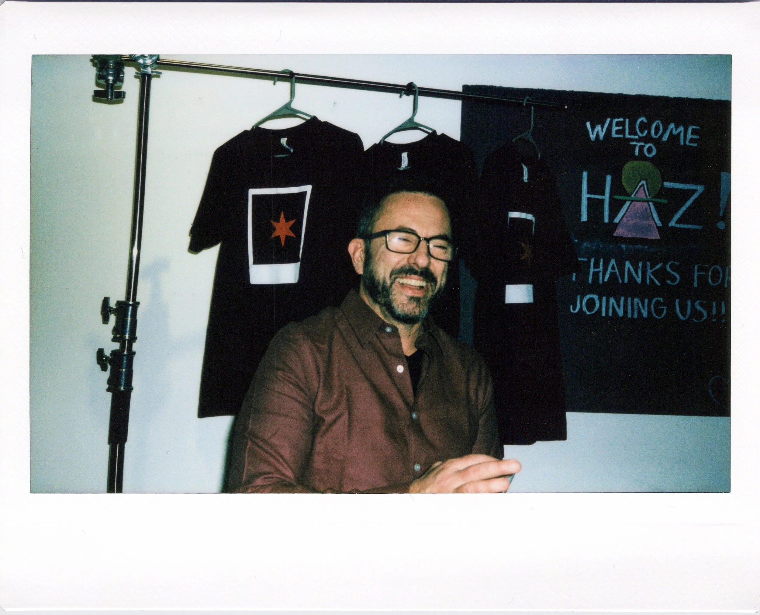 A smiling man with glasses and a beard, wearing a brown shirt, standing in front of black T-shirts with a star graphic and a blue welcome sign that says "WELCOME TO HAZ! THANKS FOR JOINING US!"
