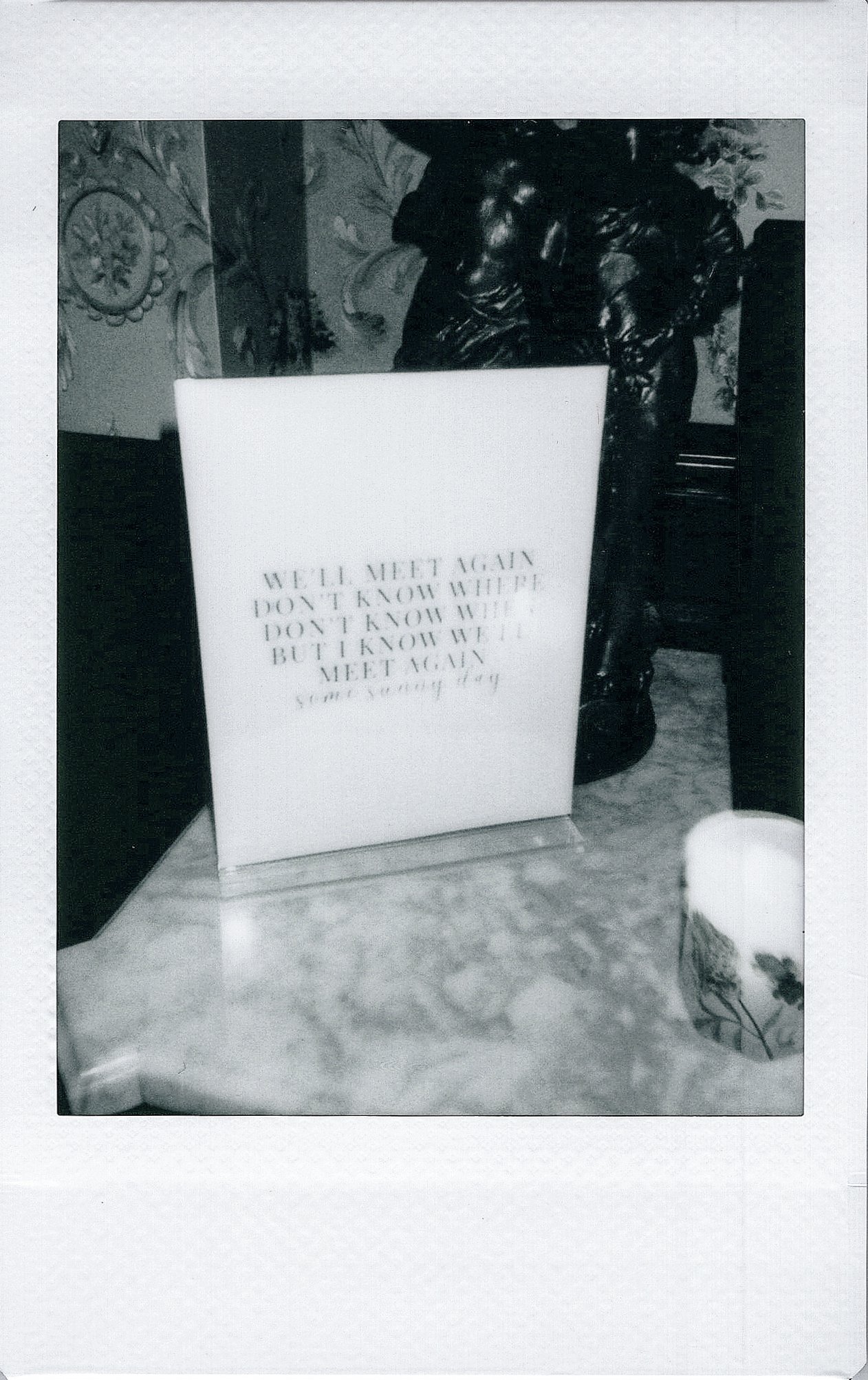 Black and white photo of a decorative statue with a sign that reads 'HELD MEET AGAIN DON'T KNOW WHERE'.