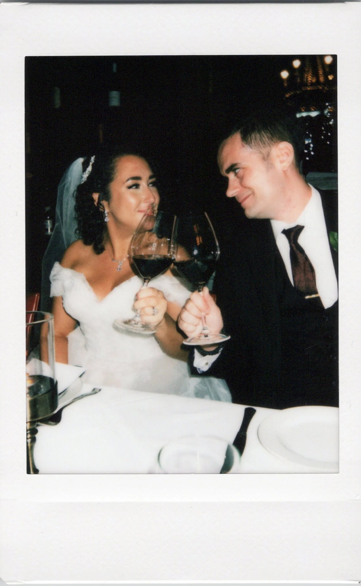 A bride and groom at a wedding reception clinking glasses of red wine, smiling and looking at each other.