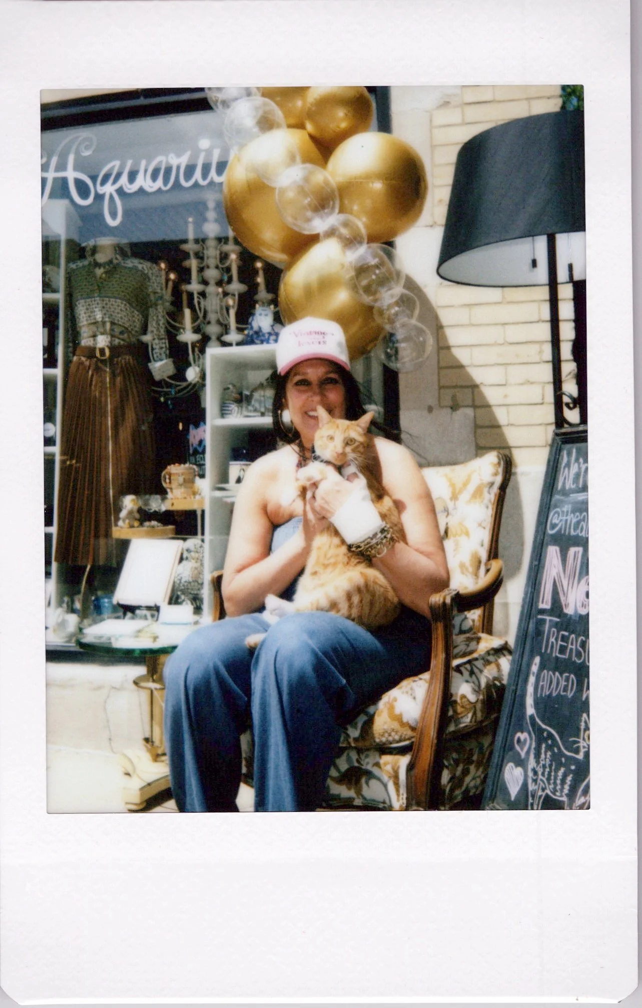 A woman sitting on a vintage armchair, holding an orange tabby cat, wearing a pink hat and smiling, with balloons and store display in the background.