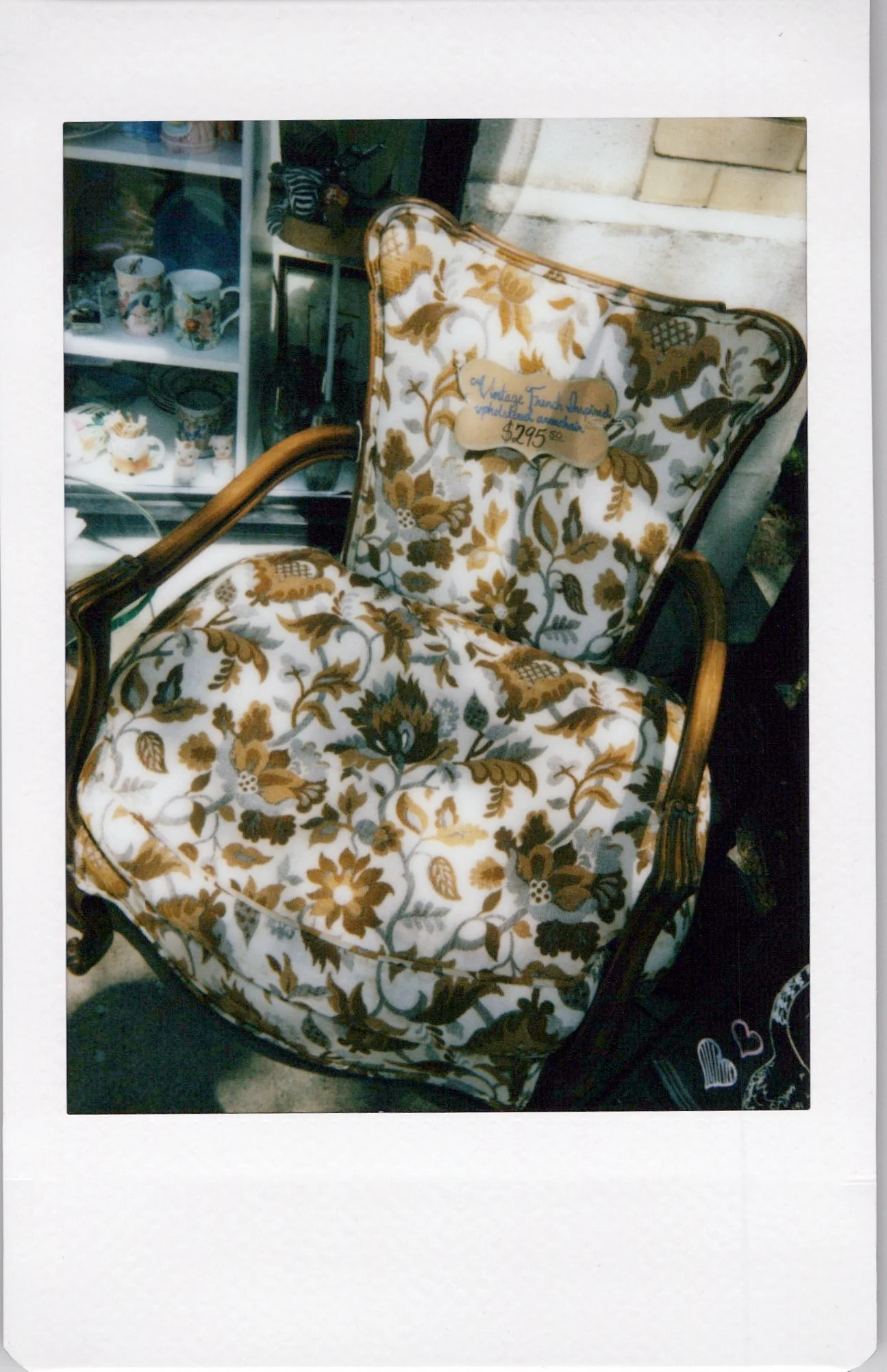 Vintage floral upholstered armchair with carved wooden frame and curved armrests, with a price tag reading $29.50. In the background, a display shelf with ceramic cups and other decorative items.
