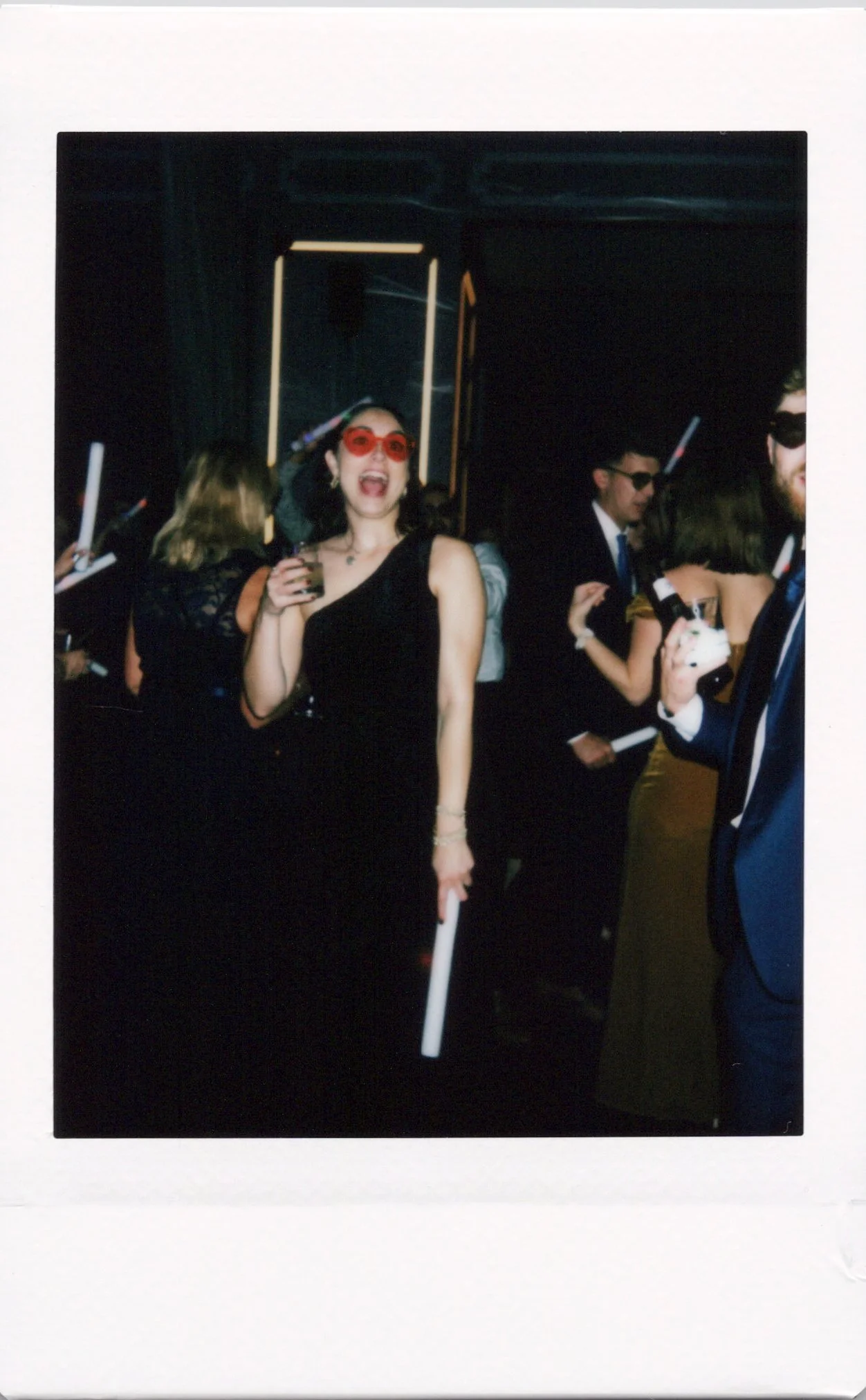 Polaroid of a group of people at a party, some wearing sunglasses, holding drinks, and dressed in formal attire, enjoying an event with a lively atmosphere.