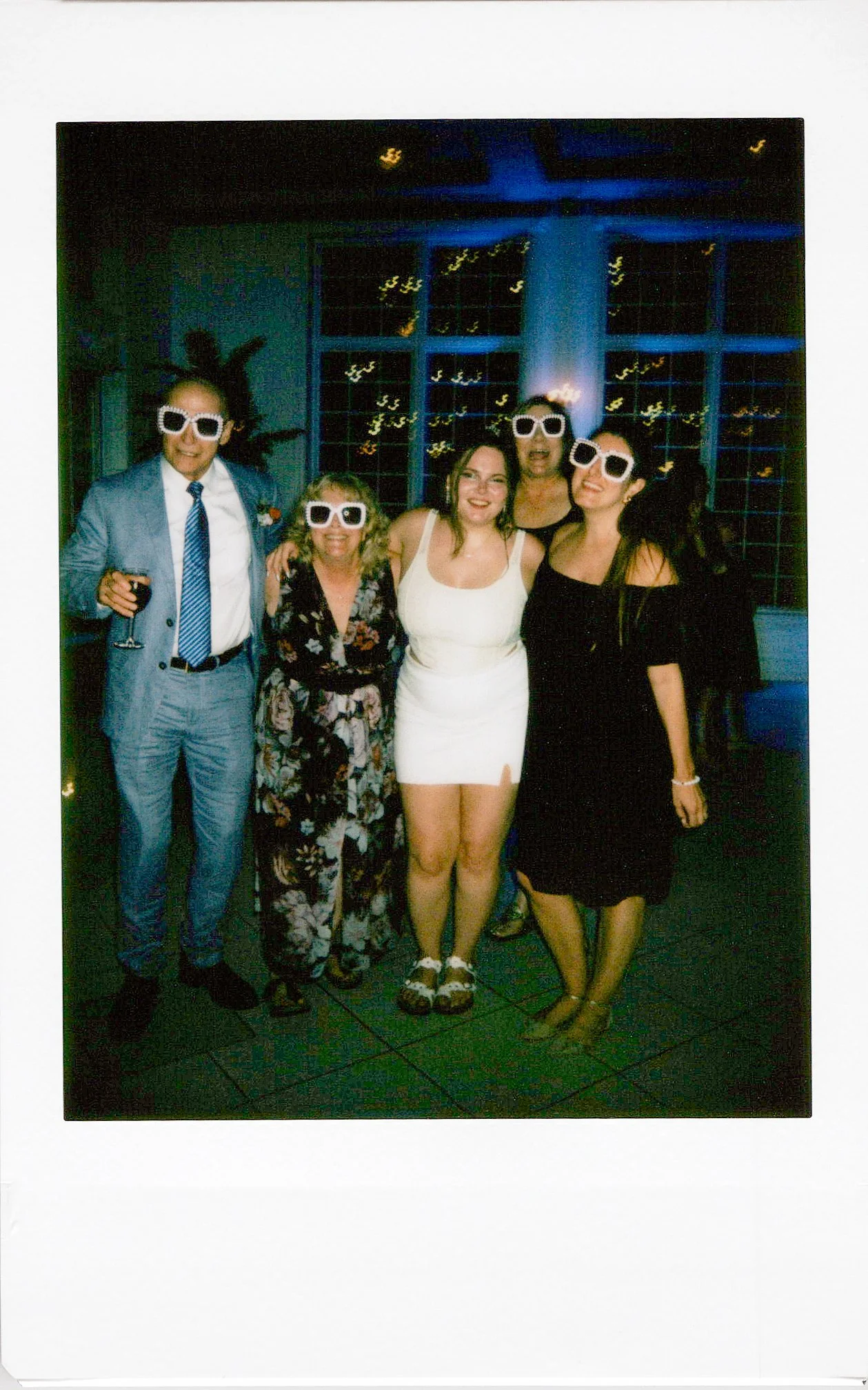 Group of five people wearing large white sunglasses, standing together indoors in front of a dark background with large window panes and city lights visible through the windows.